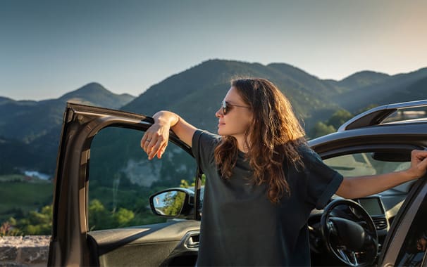 A woman stands next to her car, and she is leaning on the door. There is a picturesque mountain scene behind her.