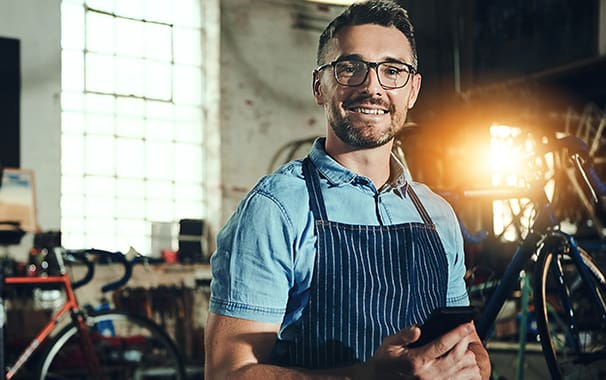 A man wearing a navy blue apron over his blue short sleeve shirt. He is standing in a shop, and he is looking at the camera and smiling.