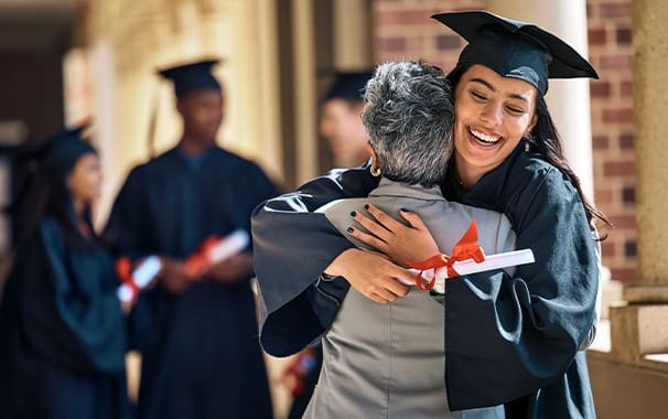 A young woman in a graduation cap and gown hugs an older woman. The graduate is holding a diploma in her hand.