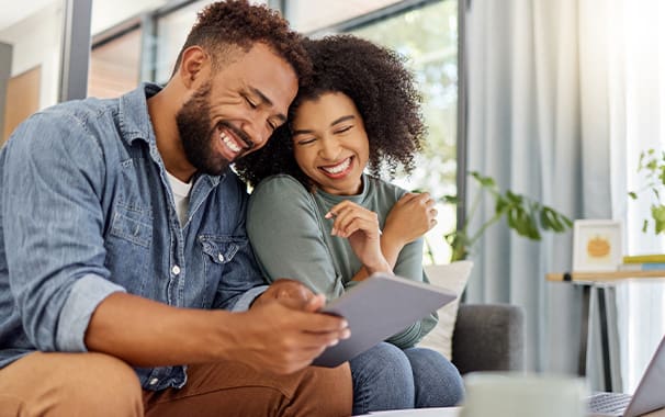 A man and a woman are seated next to each other on a sofa. He is holding a tablet and they are both looking at it and smiling