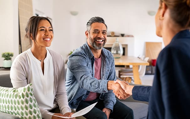 A man and woman are seated next to each other and smiling. The man's arm is outstretched shaking the hand of someone who is not pictured