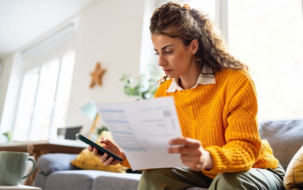 A woman is holding paperwork in her left hand, while looking down at the smart phone in her right hand